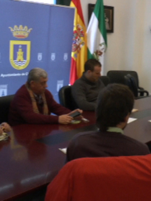 Sala de conferencias con una mesa central y sillas alrededor. En la pared, paneles informativos y banderas nacionales. Personas sentadas en sillas, algunas con teléfonos. En la pared, un proyector y pantallas de información.
