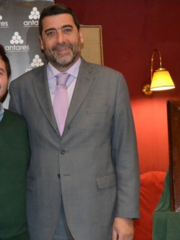 Dos hombres sonrientes posan junto a un premio en una sala de conferencias. El hombre izquierdo lleva una camisa verde y el derecho, un traje gris con corbata rosa. Detrás de ellos hay una pantalla con el logo de Antares y un cartel informativo sobre "Kike Esprosa". La imagen se toma en una sala con un fondo rojo y una lámpara de pie.