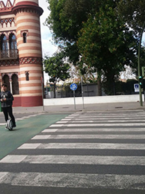 Se puede observar una calle con un paso peatonal en el centro, frente a un edificio de estilo colonial con torres rojas y blancas. El cielo es claro y hay árboles al fondo, lo que sugiere un entorno urbano.