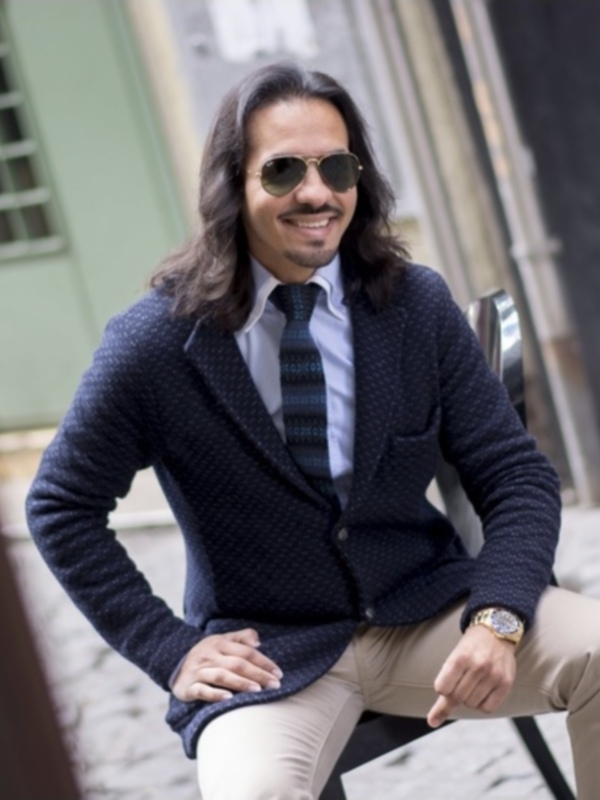 Un hombre con cabello largo y gafas de sol se sienta en una calle adoquinada, vestido con un traje azul y camisa blanca. Detrás de él hay una pared verde con un ventanal.