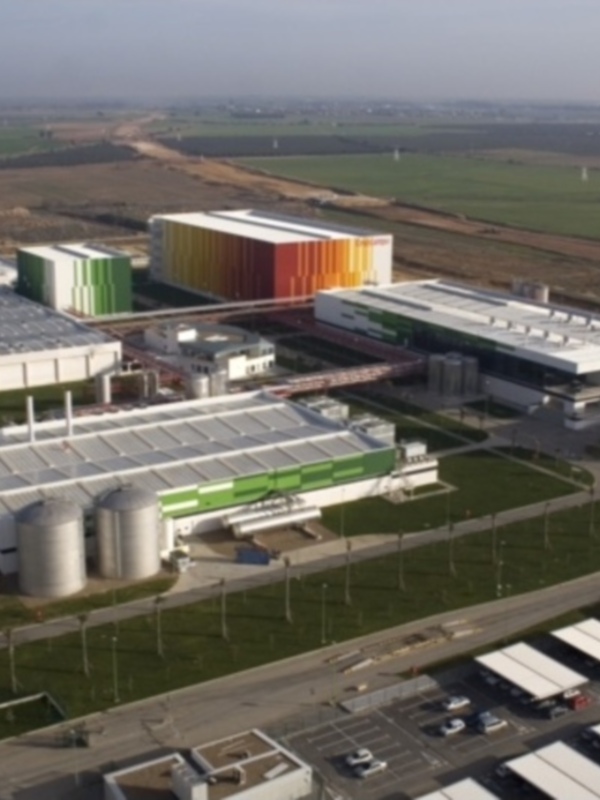 Vista aérea de una moderna fábrica industrial con edificios de colores vibrantes y paneles solares en el techo, rodeada de campos verdes y carreteras.