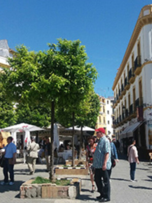 En un entorno urbano, se observa una plaza con edificios de estilo típico español. Hay personas caminando, un paraguas en el centro y una estatua en la esquina. El cielo está despejado, indicando buen tiempo.