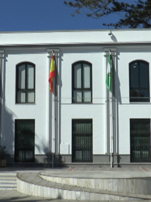 Fachada de un edificio blanco con ventanas y puertas negras, banderas en las columnas. Suelo de piedra con escaleras y árboles al lado.