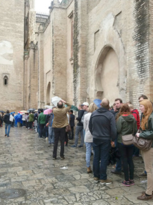Una multitud de personas en una fila, esperando frente a un edificio histórico con arcos y murallas. La gente está vestida informalmente, algunas con sombrillas para protegerse del sol. El entorno es de piedra y la arquitectura parece antigua, posiblemente un monumento o atracción turística.