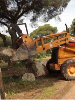 Una excavadora CASE está trabajando en un camino de tierra, arrancando grandes bloques de roca. El entorno es una zona forestal con árboles altos y verdes, lo que sugiere un trabajo de mantenimiento o construcción en una zona rural.