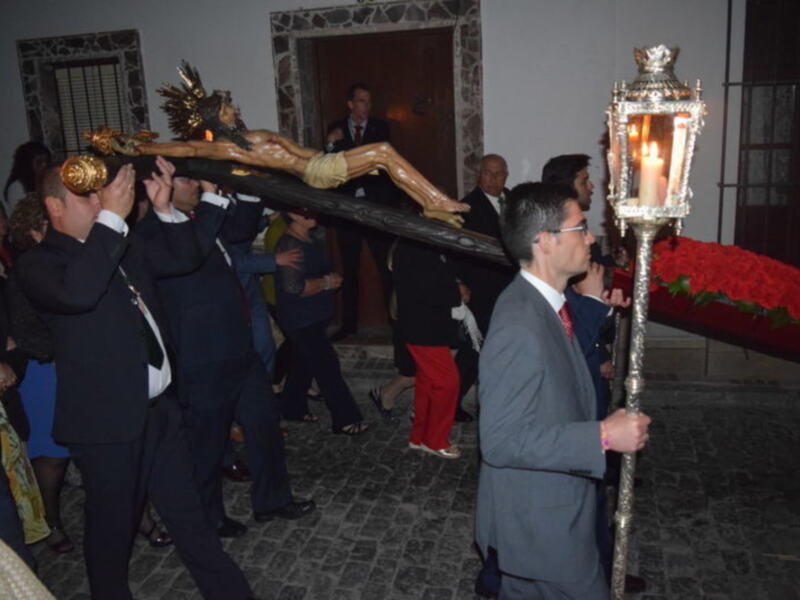 Imagen de una procesión religiosa en un entorno interior, con personas vestidas formalmente llevando un ataúd y una figura de madera. La iluminación proviene de una linterna portada por un hombre en el frente, enfocándose en la escena ceremonial.