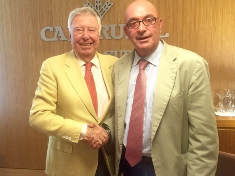 Dos hombres en trajes se saludan con una sonrisa, frente a un fondo de madera con la palabra "CAMPANA" visible.