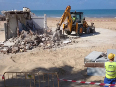 Derrumbe de una estructura en la playa, con un excavador desmantelando el edificio. El mar y el cielo azul forman el fondo. Un trabajador con un chaleco amarillo supervisa el proceso. Se han colocado barreras de seguridad alrededor del área de trabajo.