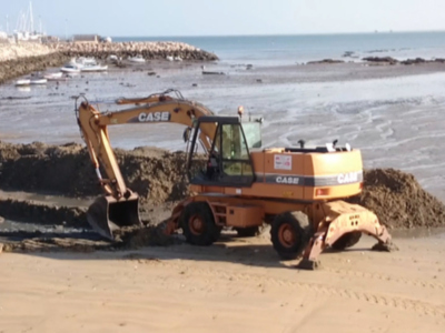 Excavadora amarilla trabajando en la orilla del mar, con agua y rocas al fondo.