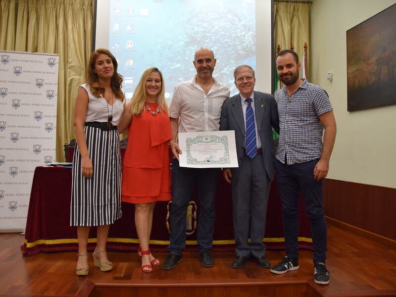Una imagen de una ceremonia de premiación en un ambiente formal. En el centro, una persona sostiene un diploma mientras está rodeada por cuatro personas que la saludan. El fondo muestra una pantalla con logotipos y un retrato enmarcado, junto a cortinas amarillas. La escena transmite un ambiente de celebración y reconocimiento.