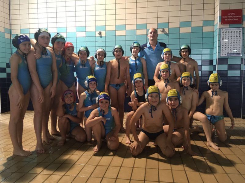 Niños de una escuela de natación posando en el fondo de una piscina, todos con trajes de baño y gorros de natación, con un instructor en el centro. La pared de la piscina está decorada con azulejos azules y blancos.