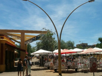 Un parque con sombrillas y mesas, gente caminando, árboles y un cielo azul.