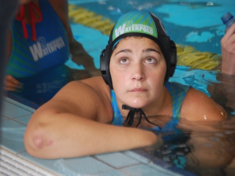 Una nadadora con un casco verde y azul en una piscina. La imagen muestra a la nadadora con su cuerpo parcialmente sumergido en el agua, con una mano levantada. La piscina tiene azulejos blancos y azules, y hay una persona en el fondo con un traje de baño azul.