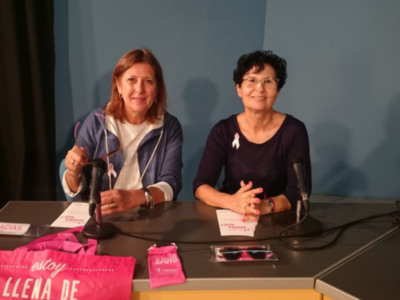 Dos mujeres sentadas frente a una mesa con pancartas y bolsas rosa, probablemente en un evento o conferencia sobre salud mental.
