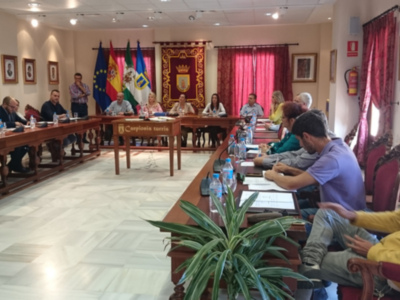 Sala de reuniones con banderas y un escudo en el fondo. Personas sentadas en mesas, hablando y escribiendo. En la mesa hay botellas de agua y documentos. La sala tiene una decoración formal con cortinas rojas y un suelo de mármol.