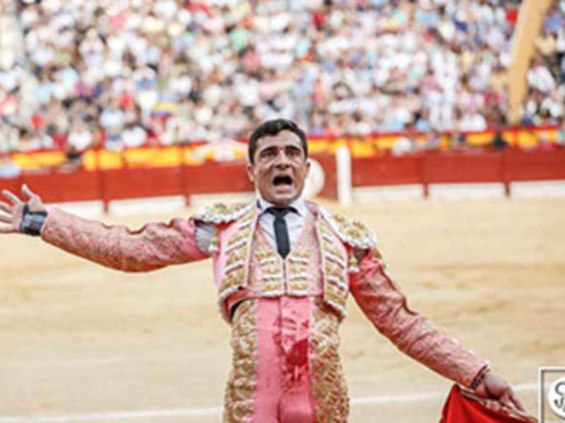 Un torero en traje de luces, con el rostro cubierto por una máscara tradicional española, realiza un gesto de triunfo con las manos extendidas. La plaza está llena de espectadores, y la arena es claramente visible en el fondo.