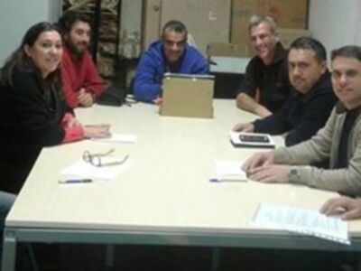 Una reunión de trabajo en un entorno laboral, con personas sentadas alrededor de una mesa, sosteniendo documentos y dispositivos electrónicos. La atmósfera es profesional y concentrada.