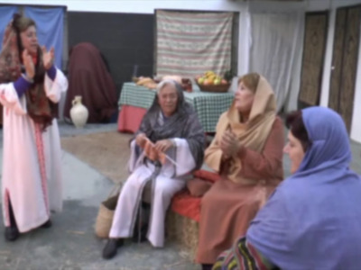 Tres mujeres en trajes tradicionales conversan en un entorno cultural, con una mesa de frutas y una tela a la pared.