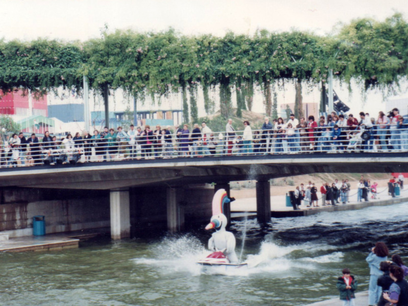 Una imagen de un puente sobre un río con una figura flotante en el agua. La gente está observando la escena, y hay un edificio rojo con una bandera en el fondo. La imagen parece ser de hace tiempo, posiblemente 1980-1990.