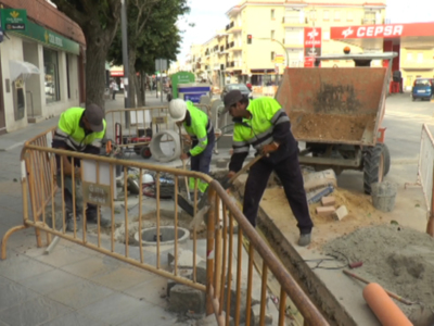 Trabajadores en verde trabajando en una calle, con un camión de volteo y una construcción en proceso.