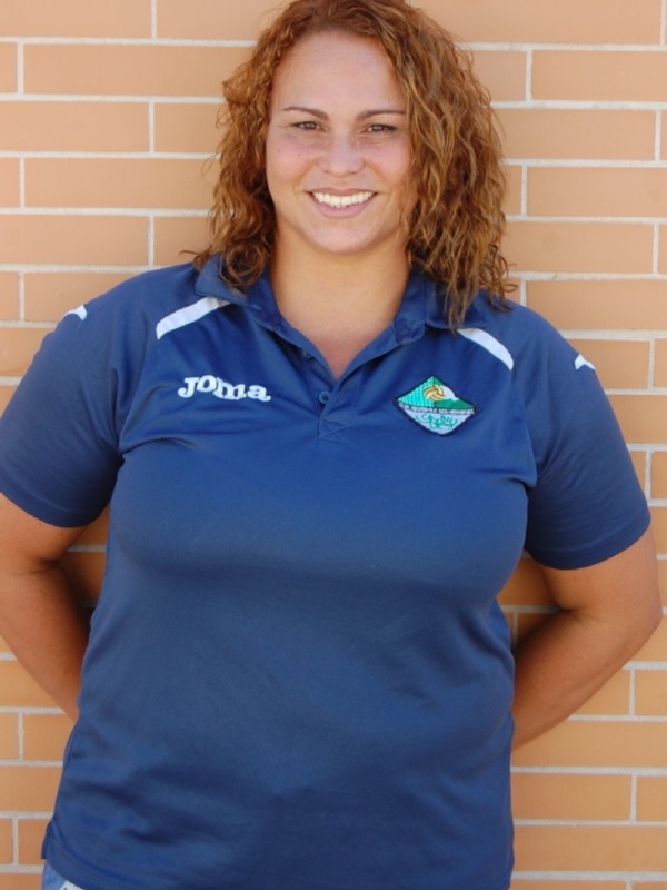 A una persona con cabello rojo, sonriendo y posando frente a un muro de ladrillo. Se puede ver parte de una camiseta azul con el nombre "Jovia" en letras blancas y un logo verde. La persona lleva pantalones azules ajustados.