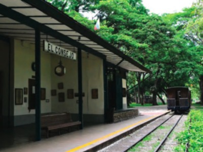 Estación histórica El Consejo, con una plataforma de madera y un andén de ferrocarril. La estación está rodeada por árboles y tiene un cartel con el nombre de la estación. La plataforma cuenta con una silla para descansar y un reloj de pared para medir el tiempo.