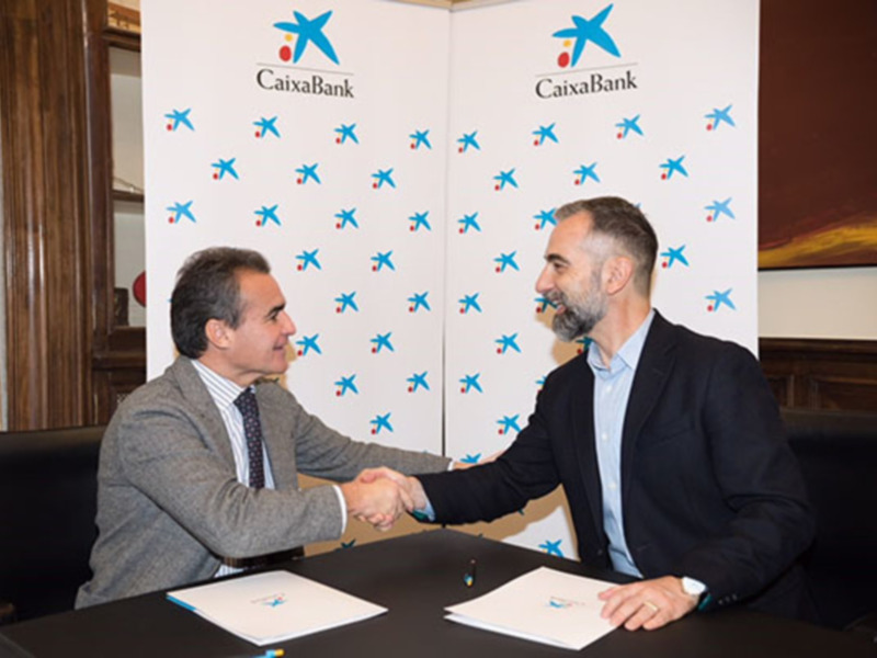 Dos hombres se saludan con un apretón de manos en una mesa, frente a un fondo con el logo de CaixaBank.