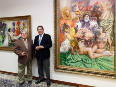 Dos hombres posan en una sala de arte, uno frente a un cuadro con figuras antiguas y el otro frente a una pintura moderna. El suelo está cubierto por un tapete de colores vibrantes, y la pared al fondo es blanca.
