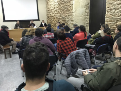Una audiencia en una sala de conferencias con un hombre en el centro, sosteniendo un teléfono móvil. La pared de fondo es de piedra y hay una pantalla en el lado izquierdo. Los participantes están sentados en sillas de diferentes colores, incluyendo azul y gris.