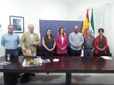 Reunión oficial en una sala con banderas españolas y europeas, posando junto a un escritorio con documentos y folletos.