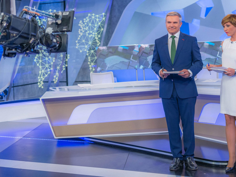 En un estudio de televisión, dos presentadores se encuentran en el centro del set. El hombre lleva un traje azul y una corbata verde, mientras que la mujer está vestida con un vestido blanco. El fondo es una combinación de pantallas y gráficos digitales que muestran información visual.