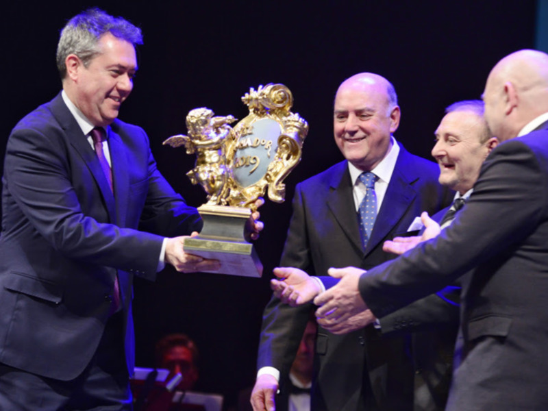 Un hombre en traje azul está entregando un trofeo dorado a otro hombre también en traje. Ambos son sonrientes y están en un escenario con luces de fondo.