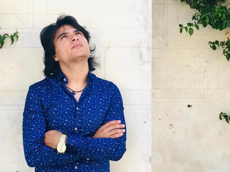 Un hombre con cabello largo y negro, vistiendo una camisa azul con pequeños patrones blancos, se encuentra de pie contra un muro blanco. Sus brazos están cruzados y mira hacia arriba, con una expresión serena o pensativa. La imagen también muestra un trozo de la pared con pequeñas marcas y una planta verde en el rincón superior derecho.