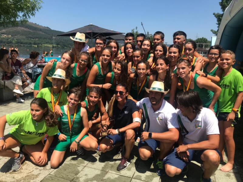 Equipo de remo celebrando con medallas y trofeos en un entorno acuático junto al lago.