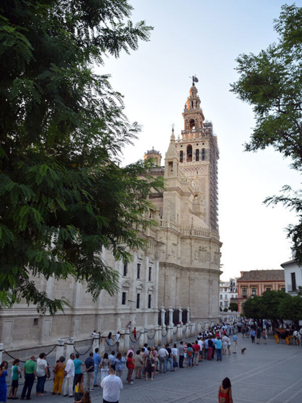 La catedral de Sevilla, una famosa iglesia gótica en España. La torre principal es visible con detalles arquitectónicos impresionantes, mientras que la plaza delante está llena de visitantes. La vista incluye árboles verdes y cielo azul, creando un entorno hermoso.