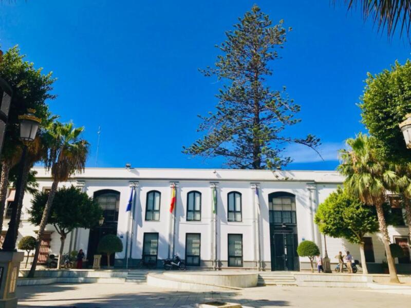 Edificio blanco con ventanas y balcones, banderas ondeando en la entrada. Árboles verdes y palmeras alrededor, cielo azul claro.