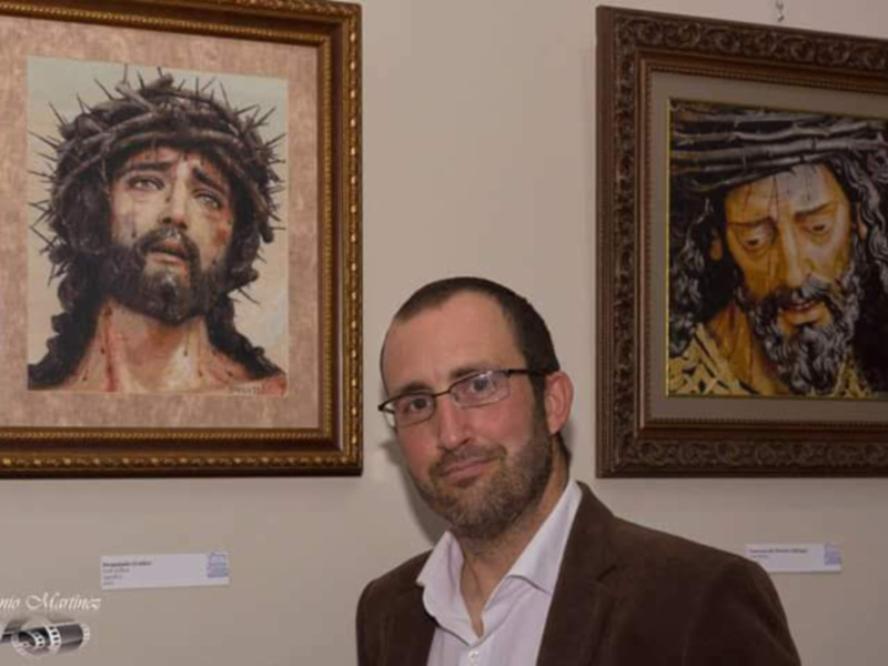 Un hombre con gafas y una camisa blanca posa frente a dos cuadros de Jesús con corona de espinas. El cuadro a su izquierda muestra un rostro más joven, mientras que el de la derecha parece ser más viejo. Ambos cuadros están enmarcados y colgados en una pared de un museo o galería.