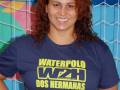 Una mujer con cabello rojo largo, sonriendo y posando frente a una valla de portería de fútbol. Lleva una camiseta azul con letras en amarillo y verde que dicen "Waterpolo DOS HERMANAS PQS". El fondo muestra un mural de colores brillantes.
