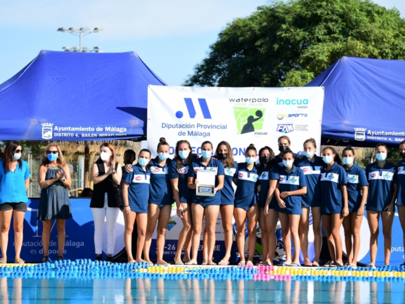 Equipo femenino de waterpolo posando frente a una piscina con banners del Ayuntamiento de Málaga y la Diputación Provincial.