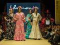 Modelos desfilan en la pasarela de moda flamenca, con vestidos tradicionales de colores vibrantes y flores en la cabeza. La escena está ambientada con un fondo de madera y un cartel que dice "Flamenca". La audiencia observa atentamente.