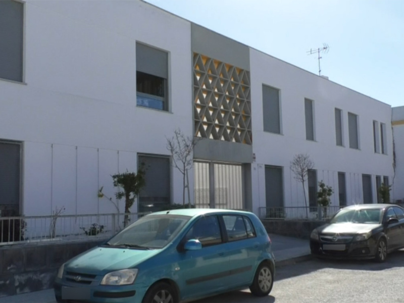 Edificio moderno con fachada blanca y diseño geométrico en la parte superior, aparcamiento en la calle con coches estacionados.