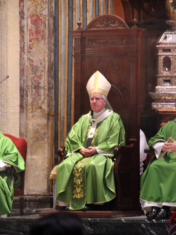 Imagen de un sacerdote vestido con un hábito verde y un casulla blanca sentado en una silla de madera. El fondo muestra detalles de la arquitectura interna de un templo con columnas y estatuas.