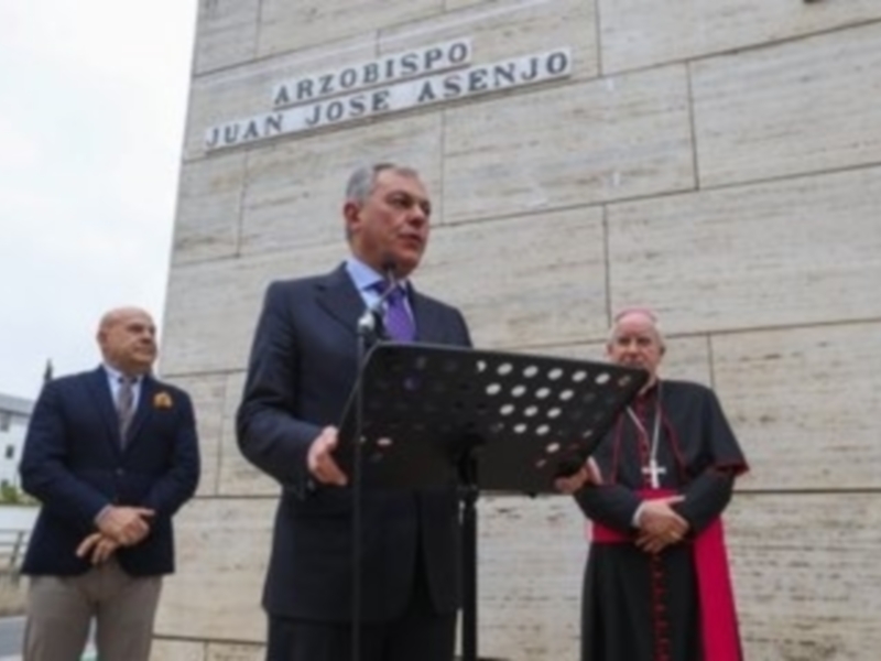 Arzobispo Juan José Asenjo hablando en un acto público frente a un monumento con su nombre en la fachada.