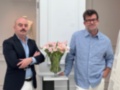 Dos hombres posan frente a una mesa con flores, uno con un traje y el otro con una camisa a rayas, en un ambiente elegante.