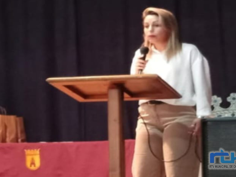 Una mujer con cabello largo y rubio, vistiendo una blusa blanca y pantalones beige, se encuentra en un escenario con un micrófono en la mano. Está frente a una mesa de madera y detrás de ella se puede ver un escudo con letras en el fondo.