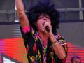 Un hombre con un afro negro y una camisa estampada de flores realiza una gesticulación con el dedo índice levantado en un escenario de concierto. La iluminación es intensa con luces rojas y amarillas en el fondo, enfocándose en la figura del cantante.