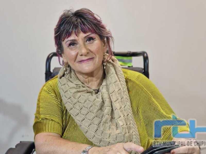 A una persona sentada en un sillón de ruedas, con cabello rojo y corto, llevando una bufanda beige. La persona sonríe amablemente mientras se sienta en el sillón de ruedas.