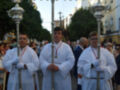 Varios hombres vestidos con trajes ceremoniales blancos, algunos con cruceros en mano, durante una procesión religiosa en una calle con edificios blancos y azules en el fondo.