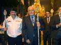 Procesión nocturna con personajes en trajes formales y ornamentos religiosos, iluminada por farolas y con una imagen procesional en el fondo.