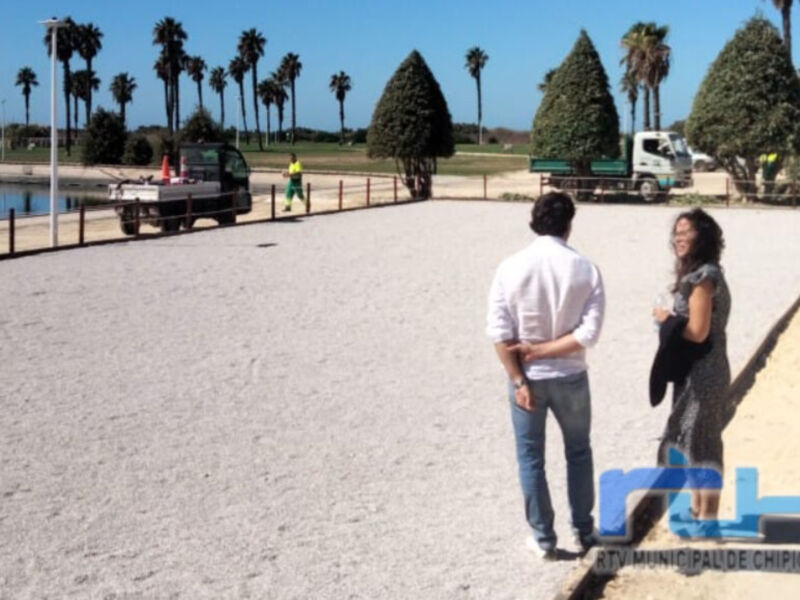 Imagen de dos personas en un área pública, con palmeras y camiones en el fondo. Logotipo de la Municipalidad de Chipiona en la esquina inferior derecha.